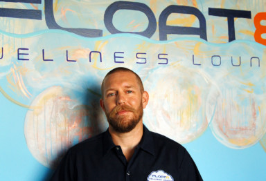 Matt Beck, owner of Float8 Wellness Lounge, standing in front of a branded mural