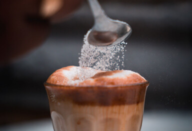 hand pouring spoonful of sugar into a frothy coffee drink