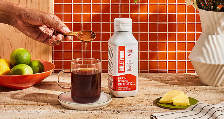 Hand pouring a spoonful of MCT oil into a cup of coffee next to a bottle of Bulletproof MCT oil