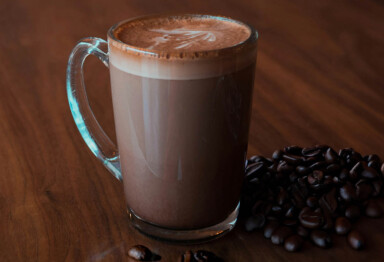 Clear mug filled with a frothy hot chocolate sitting on wooden surface and surrounded by coffee beans